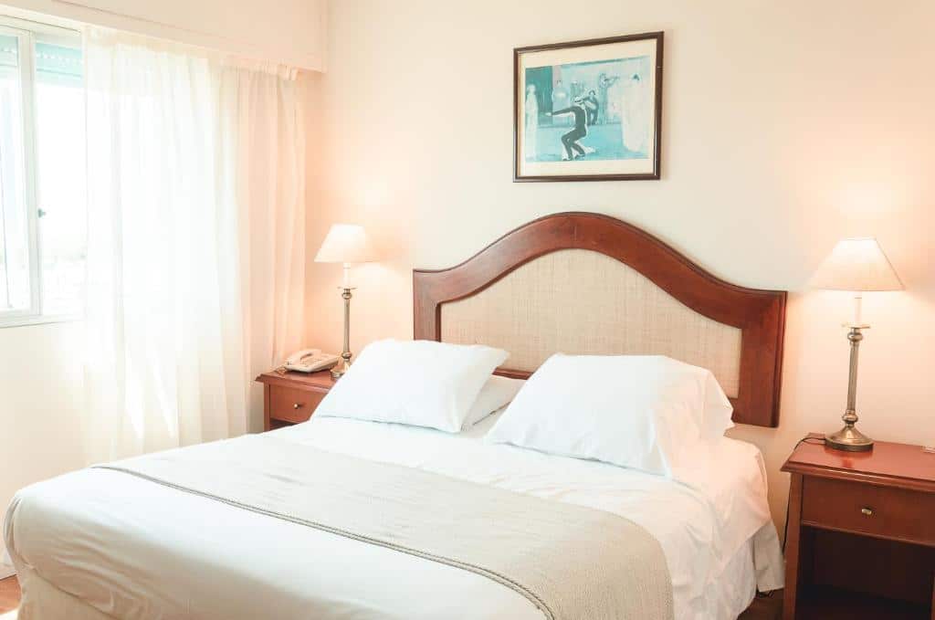 Uma cama bem arrumada com roupa de cama branca entre duas mesas de cabeceira, cada uma com uma lâmpada, sob um quadro emoldurado na parede, no Hotel Sunset Beach (Praia Mansa), para representar onde ficar em Punta Del Este.