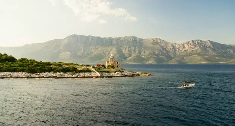 A imagem mostra uma paisagem costeira da ilha de Hvar, na Croácia. No centro da imagem, há um pequeno farol e uma casa de pedra com telhado vermelho, situados em uma península rochosa que se projeta para o mar Adriático. O farol é de cor clara e está cercado por vegetação verde. Ao fundo, há uma cadeia de montanhas altas e imponentes, criando um contraste dramático com o mar azul escuro em primeiro plano. À direita, no mar, há um pequeno barco a motor navegando, deixando uma trilha de ondas suaves na água. O céu está claro, com algumas nuvens brancas dispersas, e a luz do sol ilumina suavemente a cena.