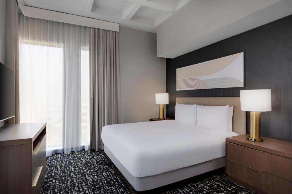 Quarto do Hyatt House LAX Century Blvs. Do lado direito uma cama de casal, uma cômoda de cada lado e um abajur. Do lado esquerdo uma raque com televisão, no fundo uma janela. Foto para ilustrar post sobre hotéis baratos em Los Angeles.
