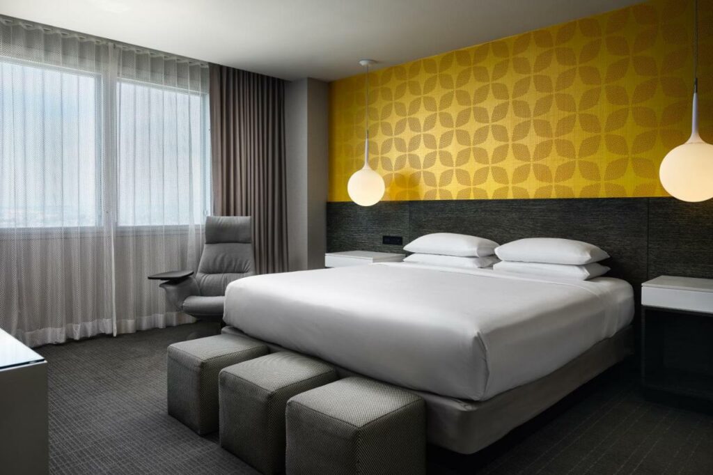 Quarto do Hyatt Regency Los Angeles International Airport. Do lado direito uma cama de casal com uma prateleira e um lustre de cada lado. Na frente três puffs. No fundo uma poltrona e uma janela. Foto para ilustrar post sobre hotéis baratos em Los Angeles.