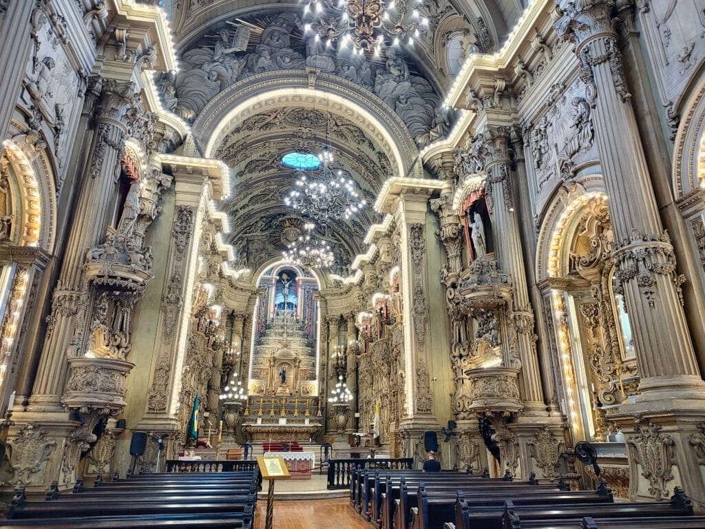 Um interior de igreja magnífico, com uma arquitetura barroca rica em detalhes. Arcos e colunas ornamentadas ladeiam o caminho até o altar, que é decorado com esculturas e entalhes intrincados. Lustres pendem do teto, iluminando o espaço com uma luz suave. O teto abobadado é adornado com relevos elaborados e pinturas, criando uma atmosfera de grandeza e reverência. Bancos de madeira escura estão dispostos em fileiras, conduzindo o olhar dos visitantes para o impressionante altar ao fundo