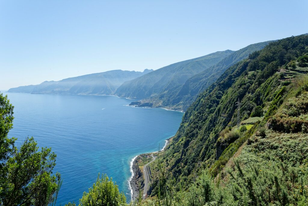 A imagem mostra uma vista panorâmica da costa da Ilha da Madeira, em Portugal. A cena é dominada por uma combinação impressionante de montanhas verdes e o mar azul profundo. À direita, uma encosta íngreme coberta por vegetação exuberante desce até a costa. A vegetação é densa e variada, com árvores e arbustos de diferentes tipos que se espalham pela montanha. No fundo, as montanhas se estendem ao longe, criando um relevo ondulado e imponente. À esquerda, o mar azul se estende até o horizonte, refletindo o céu claro. A água é calma e límpida, com tonalidades que variam do azul escuro ao turquesa, especialmente perto da costa, onde as ondas pequenas quebram suavemente.
