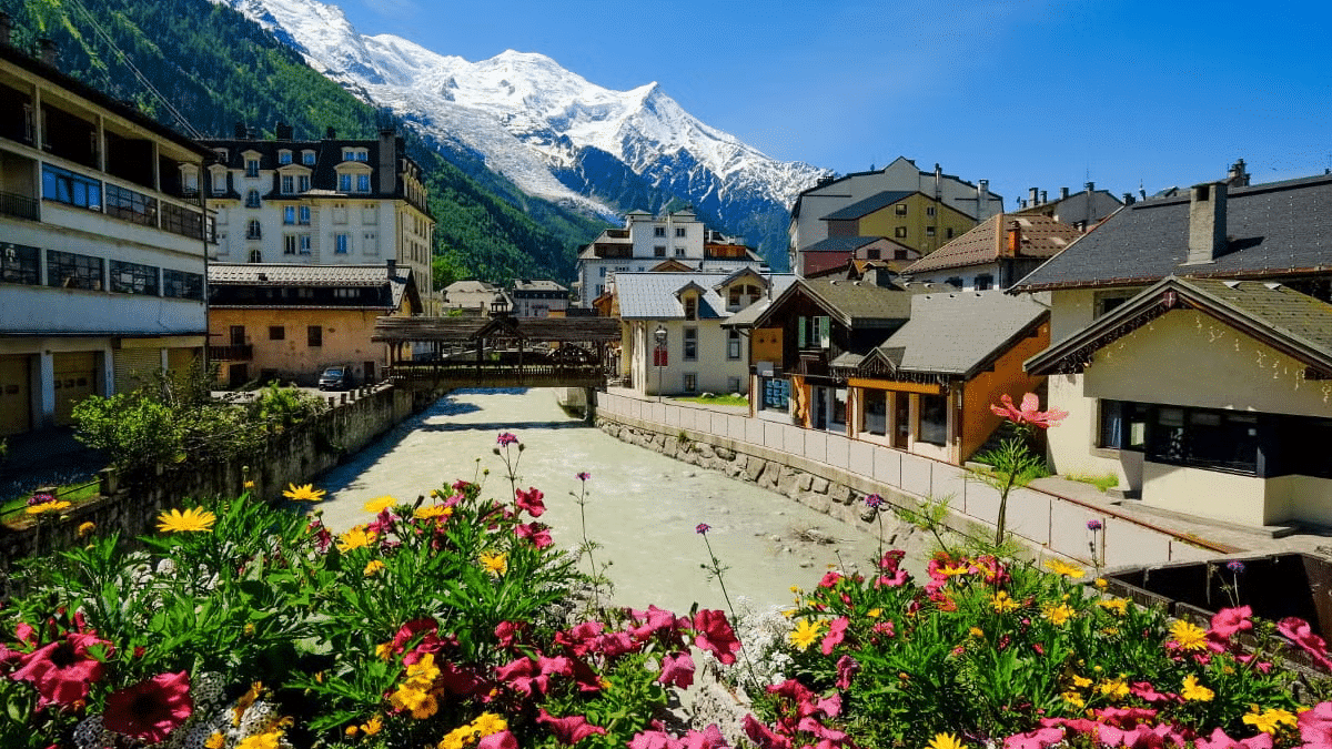 Imagem durante o dia da cidade de Chamonix com flores a frente ao fundo no centro o rio e em cada lado da imagem casas.