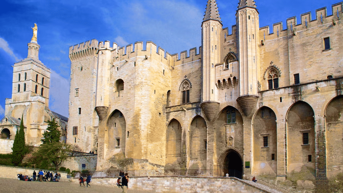 Imagem do palácio dos Papas durante o dia com pessoas andando a frente no pátio e ao fundo o edifício.