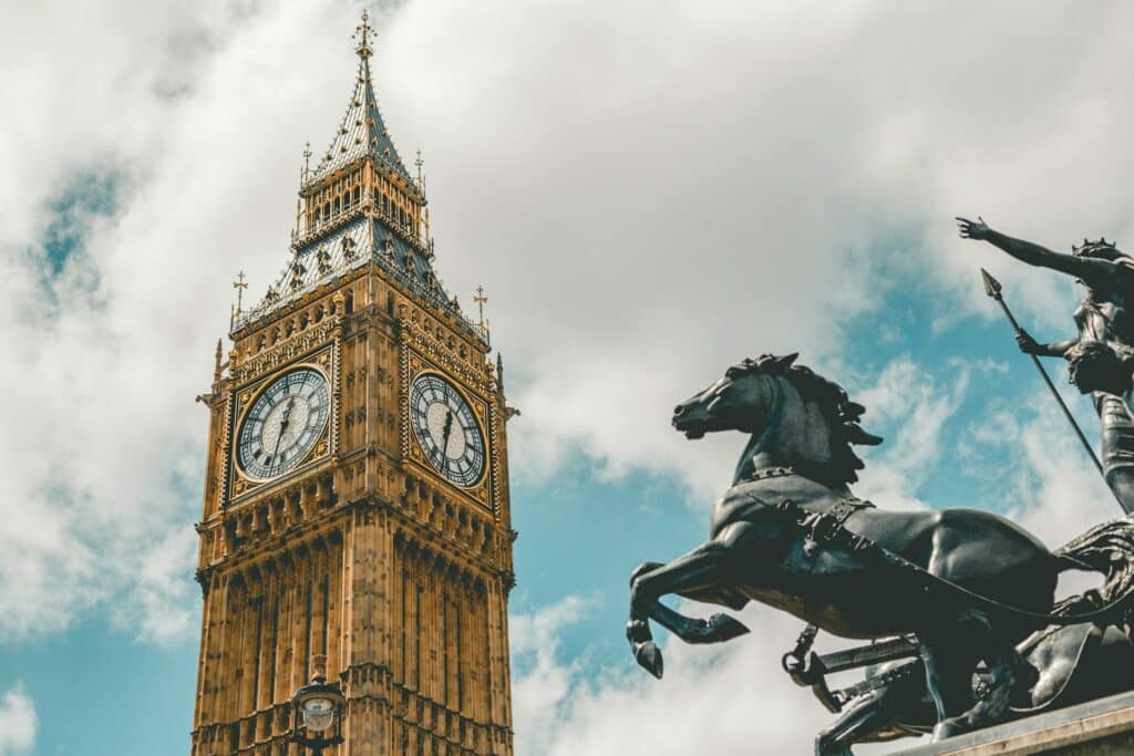 Vista debaixo do Big Ben, em Londres, Inglaterra. A torre do relógio se ergue contra o céu azul com nuvens, e ao lado há uma estátua de um cavaleiro num cavalo. Esse é um dos pontos turísticos da Inglaterra.