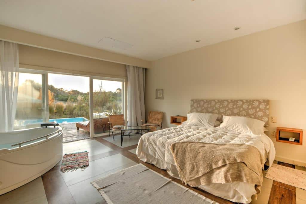 Um quarto espaçoso com uma cama de casal, cadeiras, uma mesa redonda, uma banheira de hidromassagem e portas deslizantes que se abrem para o deck da piscina, no La Viuda de José Ignacio, para representar onde ficar em Punta Del Este.