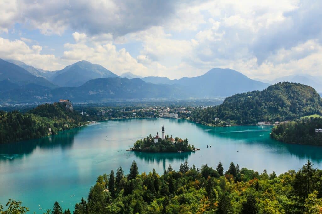 Vista panorâmica do Lago Bled na Eslovênia, famoso por sua água azul-turquesa e uma pequena ilha com uma igreja no centro. É um dos destinos incomuns da Europa. Ao fundo, montanhas cobertas por florestas e nuvens dispersas no céu completam a paisagem. O ambiente é sereno e pitoresco, destacando a beleza natural e histórica do local.
