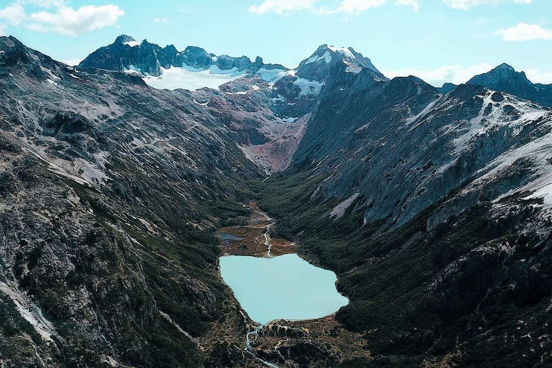 Uma vista aérea de um vale cercado por montanhas com a Laguna Esmeralda no centro. A paisagem mostra a beleza natural das formações rochosas e a vegetação ao longo do vale. Esta é uma das imagens que representam o que fazer em Ushuaia.
