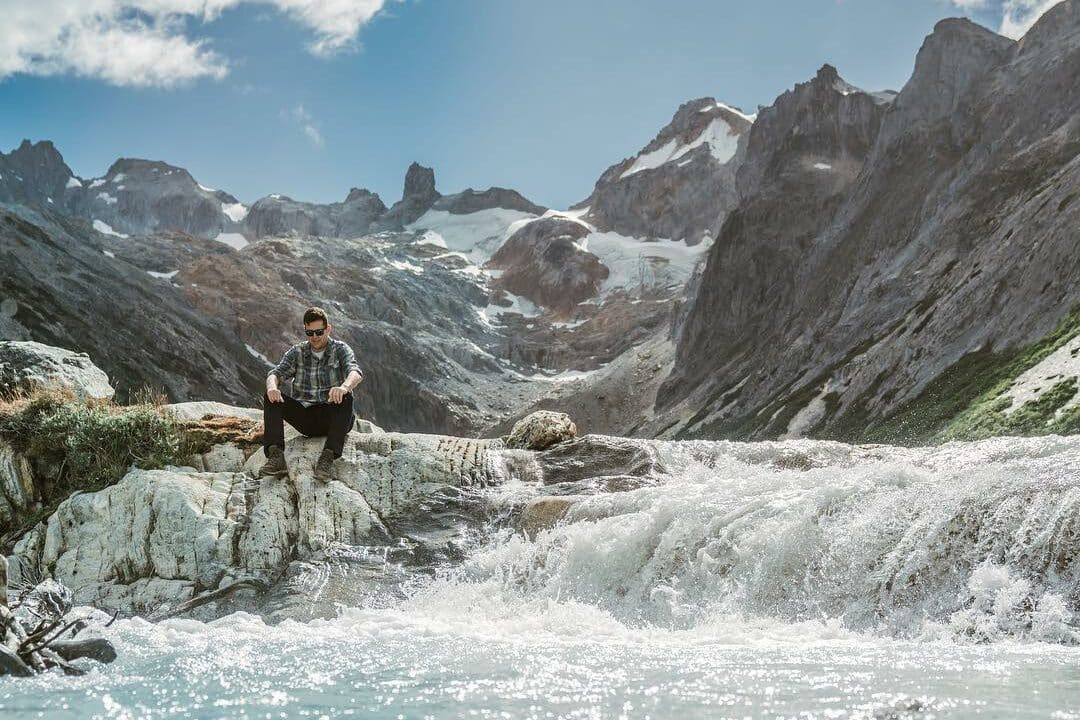 Um homem está sentado em uma rocha ao lado de uma cascata de águas cristalinas na Laguna Esmeralda, cercado por montanhas cobertas de neve ao fundo. O céu azul e algumas nuvens brancas completam o ambiente sereno.