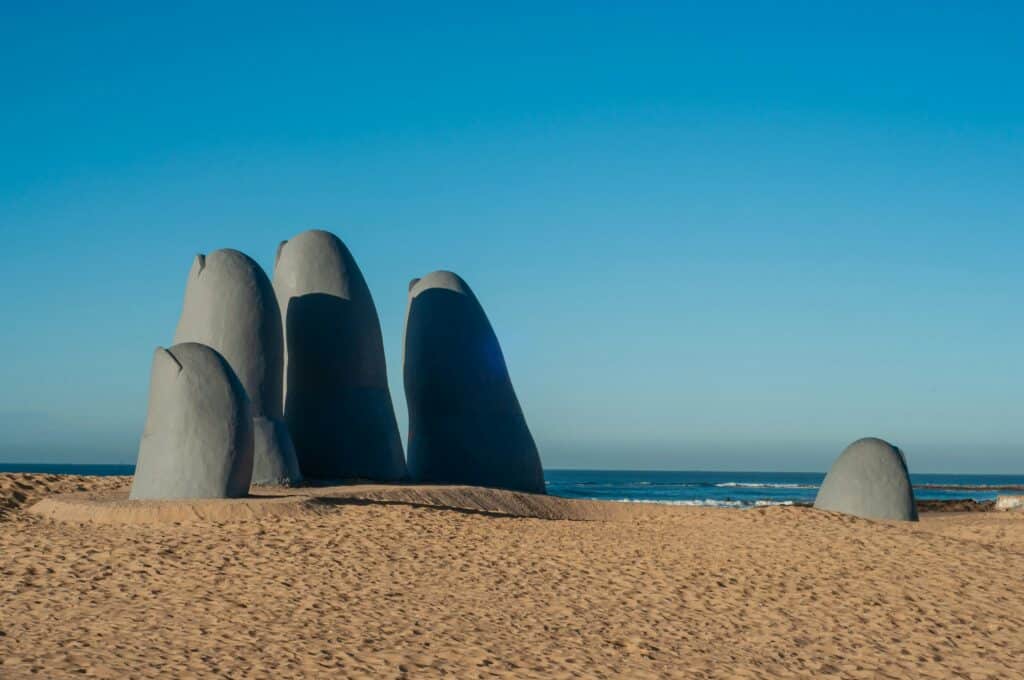 A foto está mostrando uma escultura de uma mão gigante aterrada na areia da Praia Brava, em Punta Del Este. O nome do monumento é "Los Dedos" ou "La Mano". No fundo é possível ver o mar.