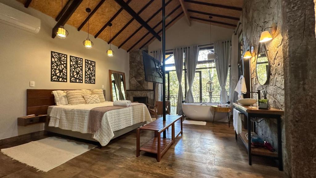 Quarto do Monte Mauá Chalés que mostra do lado esquerdo uma cama de casal, um tapete no chão, alguns quadros e espelho na parede, ar-condicionado e uma tv de frente para a cama. Já do lado direito tem uma parede de pedras com uma mesinha, dois abajures na parede e um espelho. Ao fundo do quarto tem uma banheira de hidromassagem e uma parede de vidro com cortinas que mostra a paisagem. Imagem ilustrando post O que fazer em Visconde de Mauá.