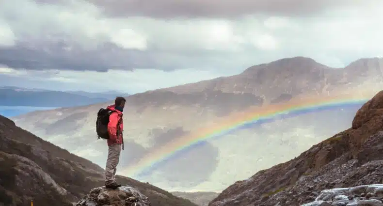 Um homem no canto esquerdo admirando um arco-íris no fundo com montanhas. Imagem para ilustrar post sobre transportes em Ushuaia.