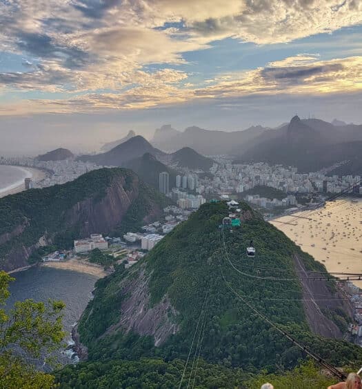 Vista aérea do Rio de Janeiro com o Pão de Açúcar, paisagem urbana e Baía de Guanabara sob um céu parcialmente nublado. Imagem para ilustrar post sobre o que fazer no feriado de Corpus Christi.