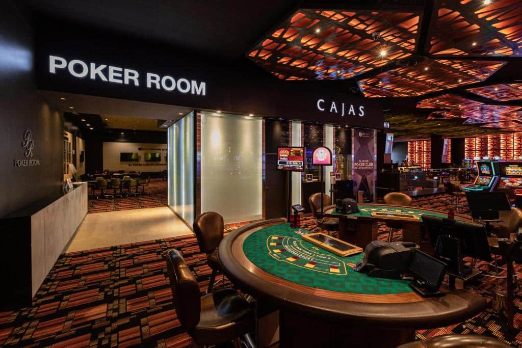 Interior de um cassino mostrando mesas de carteado, cadeiras e placas para a sala de pôquer e caixa eletrônico, com carpete estampado e iluminação, no Enjoy Punta del Este.
