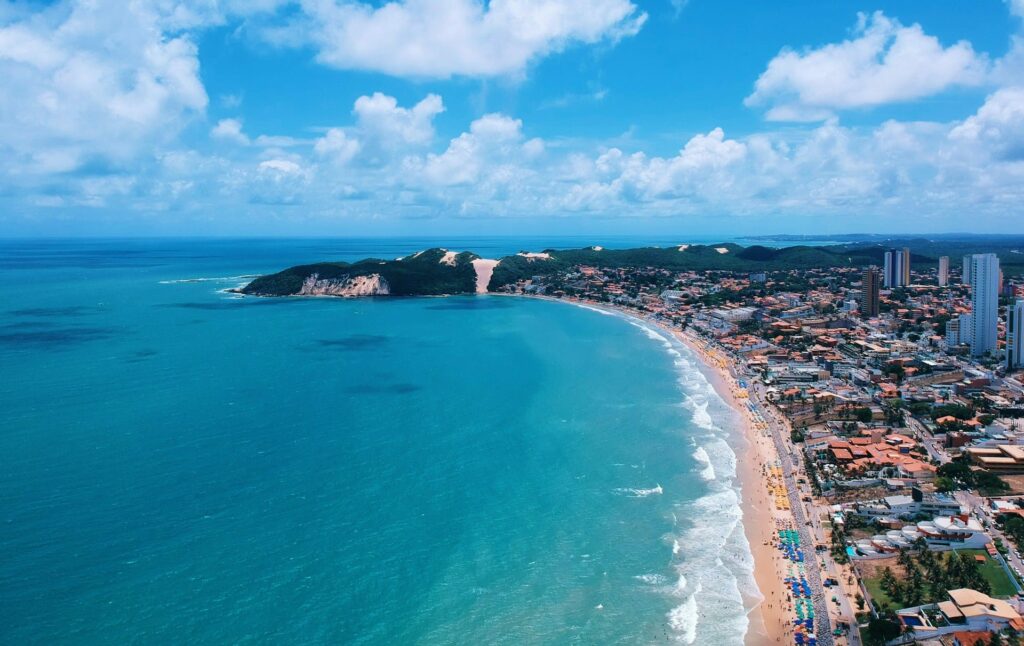 A imagem mostra uma vista aérea de Ponta Negra, uma famosa praia localizada em Natal, no estado do Rio Grande do Norte, Brasil. A praia é conhecida por seu mar azul-turquesa e suas extensas faixas de areia branca. Na linha do horizonte, é possível ver o Morro do Careca, uma duna de areia coberta parcialmente por vegetação, que é um dos cartões-postais da cidade. A praia está repleta de guarda-sóis coloridos e banhistas aproveitando o sol e o mar. O céu está claro, com poucas nuvens brancas espalhadas, e a água do mar é de um azul intenso, refletindo a luz do sol.