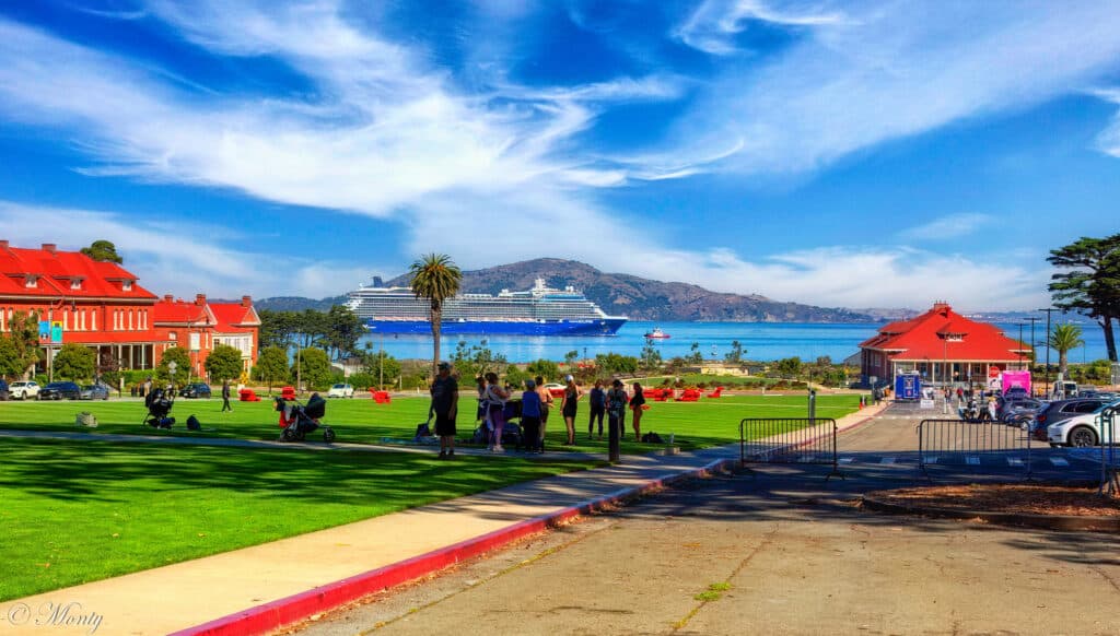 A imagem mostra uma parte do gramado e estacionamento do Parque do Presídio em San Francisco. Ao reor estão algumas construções e ao fundo está o mar e um navio.