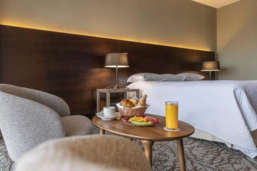 Quarto no Enjoy Punta del Este com uma cama bem arrumada, duas poltronas e uma mesa posta com café da manhã e um copo de suco de laranja, para representar onde ficar em Évora.