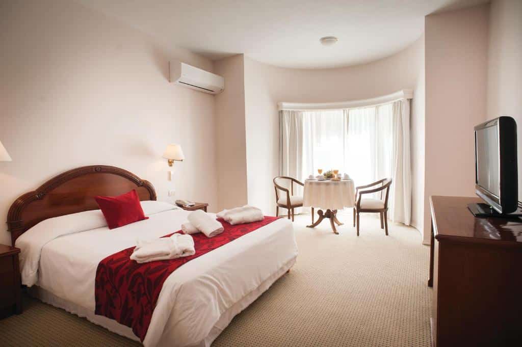 Quarto no Parque Hotel Jean Clevers com uma cama grande, detalhes em vermelho, uma TV, uma mesa com duas cadeiras perto de uma janela e um ar-condicionado na parede, para representar onde ficar em Punta Del Este.