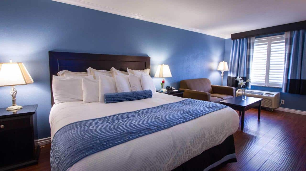 Quarto do Regency Inn in Los Angeles. Uma cama de casal do lado esquerdo com uma cômoda e um abajur de cada lado. Do lado direito uma poltrona, uma mesa de centro e uma janela.