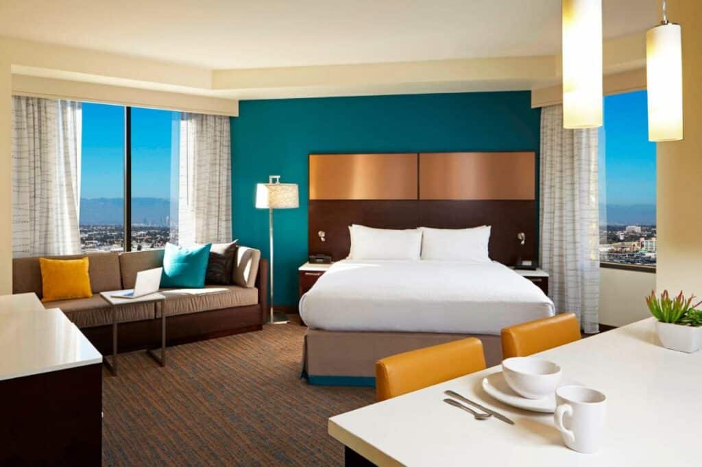 Quarto do Residence Inn by Marriott. Do lado direito uma mesa com duas cadeiras, no fundo uma cama de casal. Do lado esquerdo um sofá e uma estante. De cada lado uma janela. Foto para ilustrar post sobre hotéis baratos em Los Angeles.