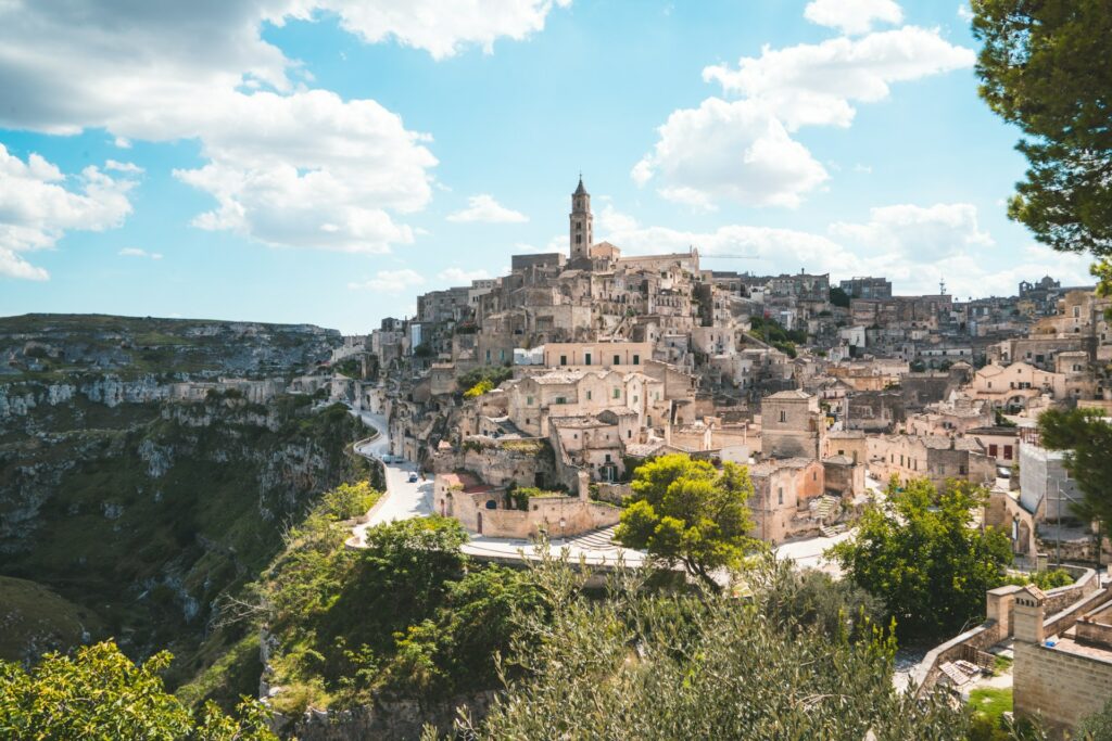 Vista de Matera, Itália, famosa por habitações escavadas na rocha, conhecidas como "Sassi". A cidade se estende ao longo de um penhasco com vista para um vale profundo. Uma igreja com uma torre alta se destaca no centro, sob um céu azul com nuvens.