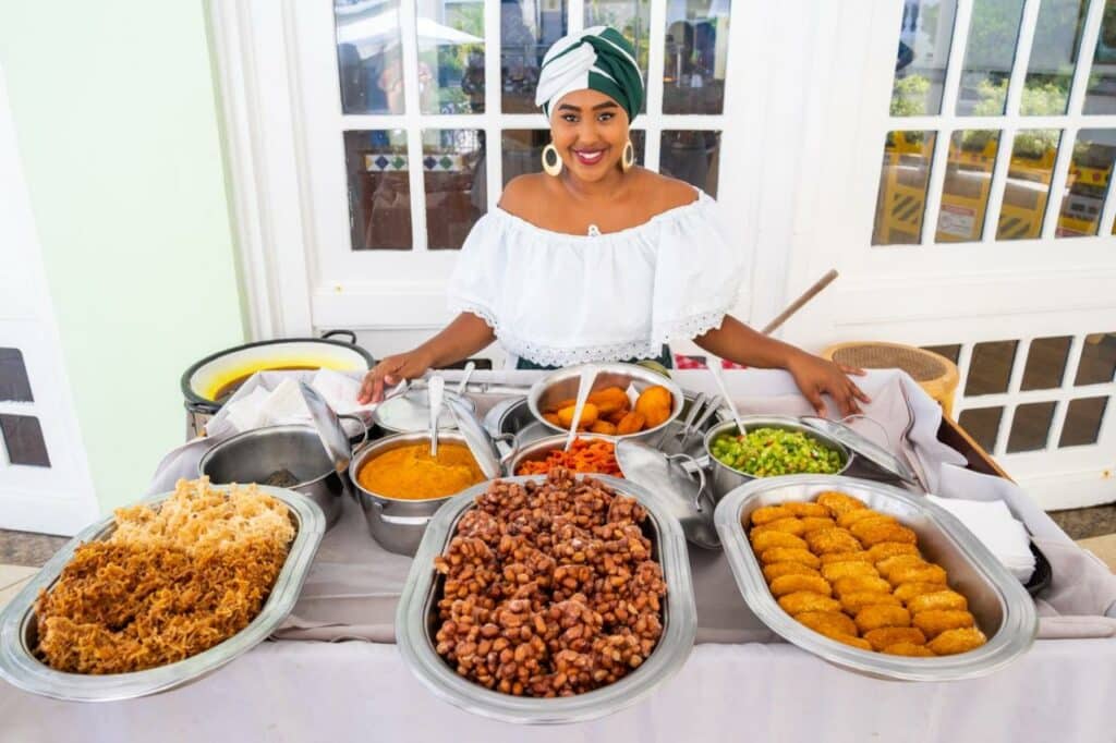 Na imagem, uma mulher sorridente, vestida com trajes tradicionais, está em frente a uma mesa cheia de comidas típicas no Sauipe Resorts Ala Terra - All Inclusive, um dos melhores resorts all inclusive no Brasil. Ela usa uma blusa branca de ombros de fora e um turbante verde e branco. A mesa está repleta de diversos pratos coloridos, incluindo feijão, farofa, acarajé e vegetais, que remetem à culinária baiana. Ao fundo, há uma porta com vidros que deixa entrever parte do ambiente externo.