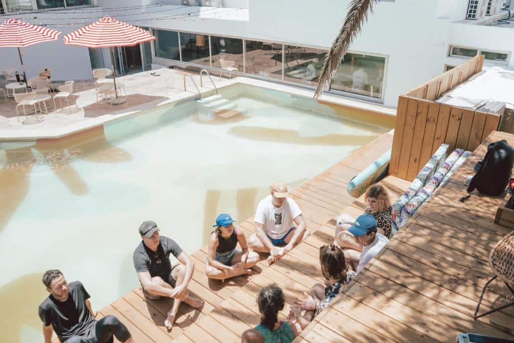 Um grupo de pessoas senta-se em círculo em um deck de madeira ao lado de uma piscina com guarda-chuvas listrados e móveis ao ar livre nas proximidades, no Selina Punta del Este.