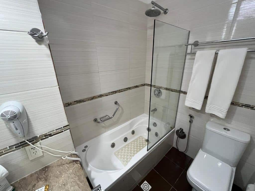 Um banheiro com banheira, chuveiro, vaso sanitário, duas toalhas brancas em um cabideiro, secador de cabelo e paredes e piso de azulejos, no Punta del Este Shelton Hotel.