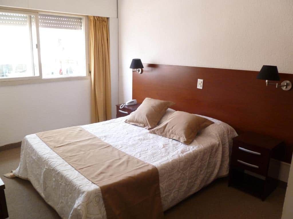 Um quarto no Punta del Este Shelton Hotel (Península) com uma cama de casal, dois travesseiros, mesas de cabeceira, luminárias de parede e uma janela com cortinas, para representar onde ficar em Punta Del Este.