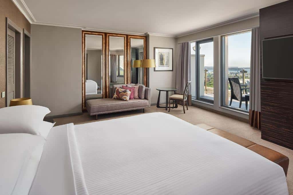 Quarto do Sheraton Grand Sydney Hyde Park. Uma cama de casal d lado esquerdo, no fundo um pequeno sofá, um armário, um abajur e uma mesa com cadeira. Do lado direito uma TV e a porta da varanda.