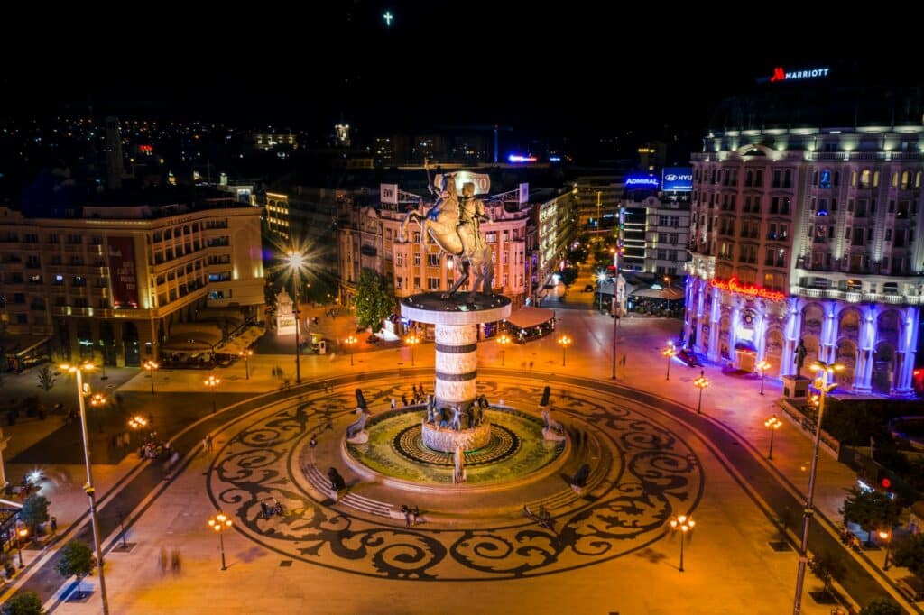Vista aérea noturna de uma praça em Skopje, a capital da Macedônia do Norte, um dos destinos incomuns da Europa. O foco é uma grande estátua no centro da praça, iluminada e cercada por padrões circulares no chão. As ruas ao redor estão repletas de luzes, e edifícios modernos e históricos iluminados bordam a cena.