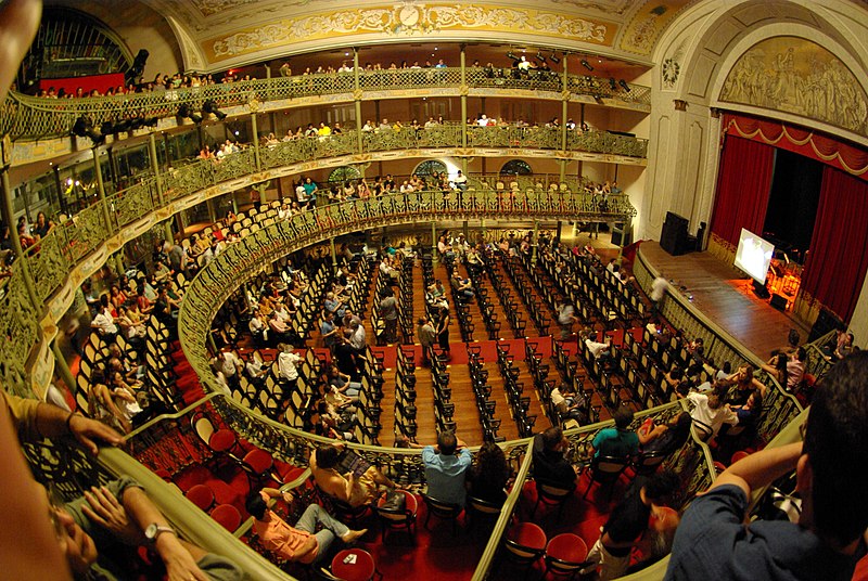 Foto tirada do interior do Teatro José de Alencar, em Fortaleza. Temos a visão de um ângulo superior, e o palco está na direita, lá embaixo. Há pessoas sentadas em assentos aleatórios, e não são todos deles que estão ocupados. O lugar é elegante, com piso de madeira e poltronas e cortinas vermelhas. Os detalhes da arquitetura são dourados e beges.