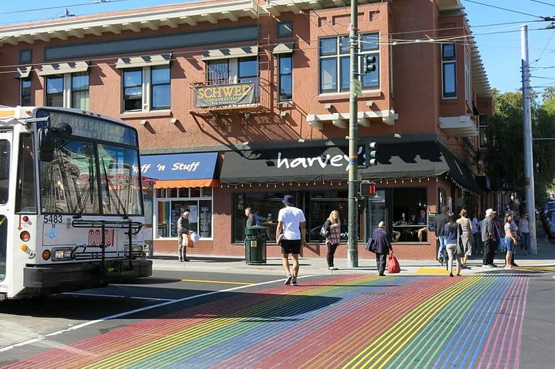 Faixa de pedrestes com as cores da bandeira LGBT+ no bairro Castro. Um ônibus está parado antes dela. Ao fundo está um prédio.