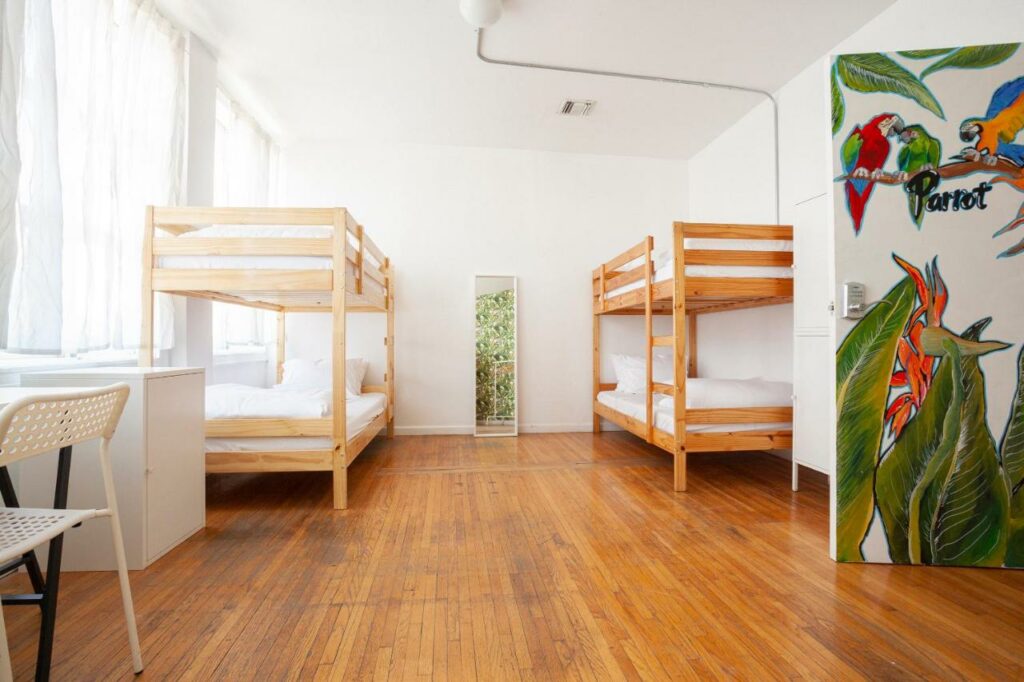 Quarto do The Steady. Do lado esquerdo janelas, na frente uma cama, uma cadeira e um armário pequeno. Do lado direito, na frente, uma porta. No fundo, de cada lado uma cama beliche e no meio um espelho.