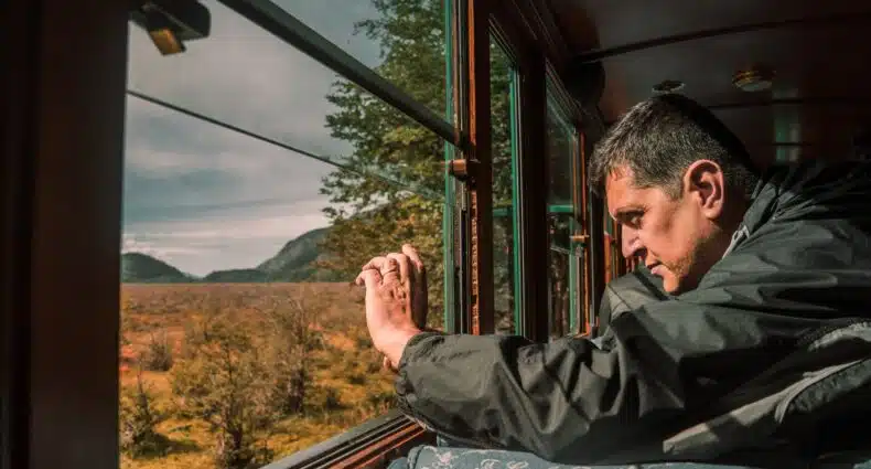 Homem branco sentado de lado, na parte direita da imagem dentro de um trem. Ele está com o braço esticado para frente, segurando um celular para fotografar a janela que mostra uma paisagem panorâmica de natureza rasteira, céus nublados e árvores verdes em Ushuaia.