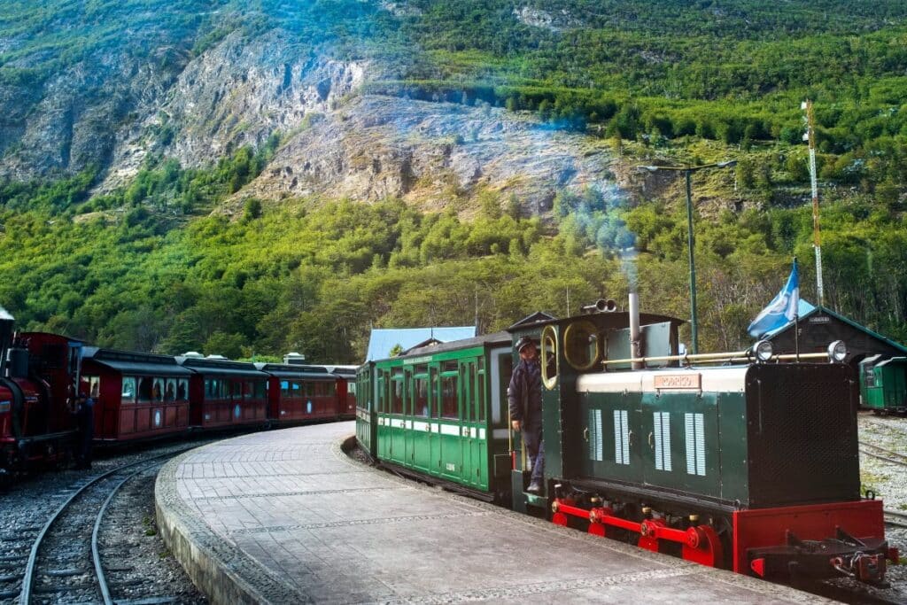 Um trem antigo de cor verde e vermelha estacionado em uma estação de trem cercada por montanhas cobertas de vegetação verde. O trem exala um pouco de fumaça, indicando que está pronto para partir.