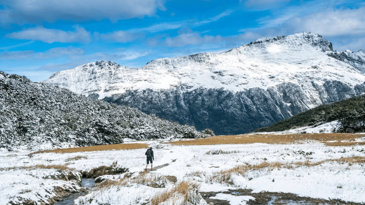 Uma trilha no meio da neve com um homem andando. No fundo montanhas.