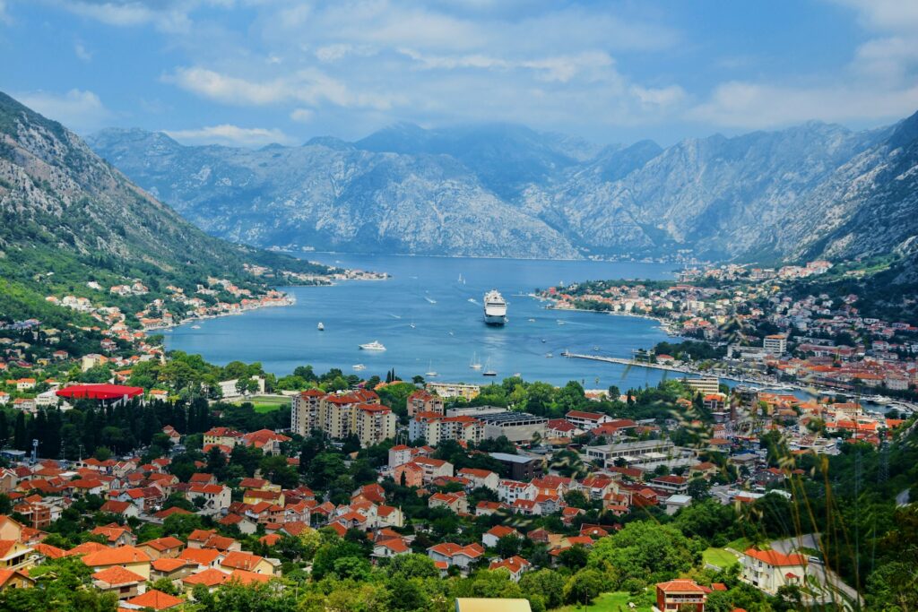 Vista panorâmica de Kotor, em Montenegro, mostrando uma baía cercada por montanhas. A cidade é composta por numerosos prédios com telhados de telha vermelha, e vegetação verde extensa. No centro da baía, um grande navio de cruzeiro está ancorado, cercado pelas águas e outras pequenas embarcações.