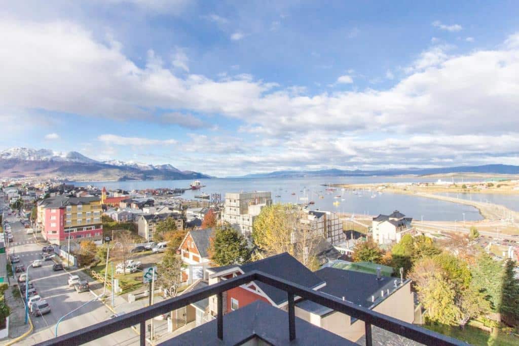 vista do centro de Ushuaia através da varanda de um dos apartamento Pacífico. Ao fundo é possível enxergar algumas montanhas com os topos nevados, além de um grande canal a frente. É possível ver as ruas da cidade e diversas construções também.