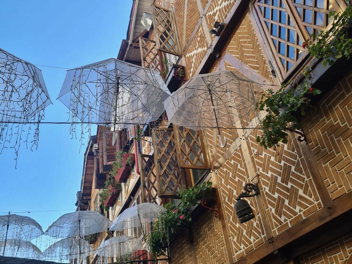 Rua entre os prédios com arquitetura suíça na Vila Capivari, onde há guarda-chuvas com piscas-piscas pendurados no ar. O céu está azul. Representa o post sobre o que fazer em Campos do Jordão.