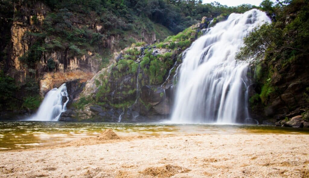 A imagem mostra a Cachoeira Maria Augusta, localizada em São João Batista da Glória, Minas Gerais. A cachoeira possui uma queda d'água significativa que desce por um paredão rochoso coberto de vegetação, formando um poço natural de água na base. O fluxo de água é volumoso e contínuo, criando uma paisagem impressionante e cheia de movimento. A água cai sobre pedras grandes e se espalha pelo leito do rio, formando pequenas corredeiras. O entorno é cercado por vegetação densa, incluindo árvores e arbustos que adicionam um toque verde à cena. Em primeiro plano, há uma faixa de areia clara que contrasta com o azul da água e o verde da vegetação, criando um ambiente natural e relaxante, ideal para um banho refrescante e contato direto com a natureza.