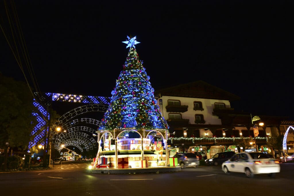 A imagem mostra uma cena noturna festiva em Gramado, Rio Grande do Sul, durante o evento Natal Luz. No centro da imagem, há uma grande árvore de Natal ricamente decorada com luzes azuis, enfeites brilhantes e uma estrela no topo. A base da árvore é adornada com presentes decorativos e luzes adicionais, criando uma atmosfera mágica.
À esquerda da árvore, a rua está iluminada com arcos de luzes decorativas que se estendem ao longo do caminho, proporcionando um ambiente festivo e acolhedor. O prédio à direita apresenta arquitetura em estilo alpino, típico de Gramado, e está decorado com luzes e guirlandas natalinas, reforçando o tema de celebração.
Há carros em movimento na rua, indicando que a área é movimentada e popular entre os visitantes.
