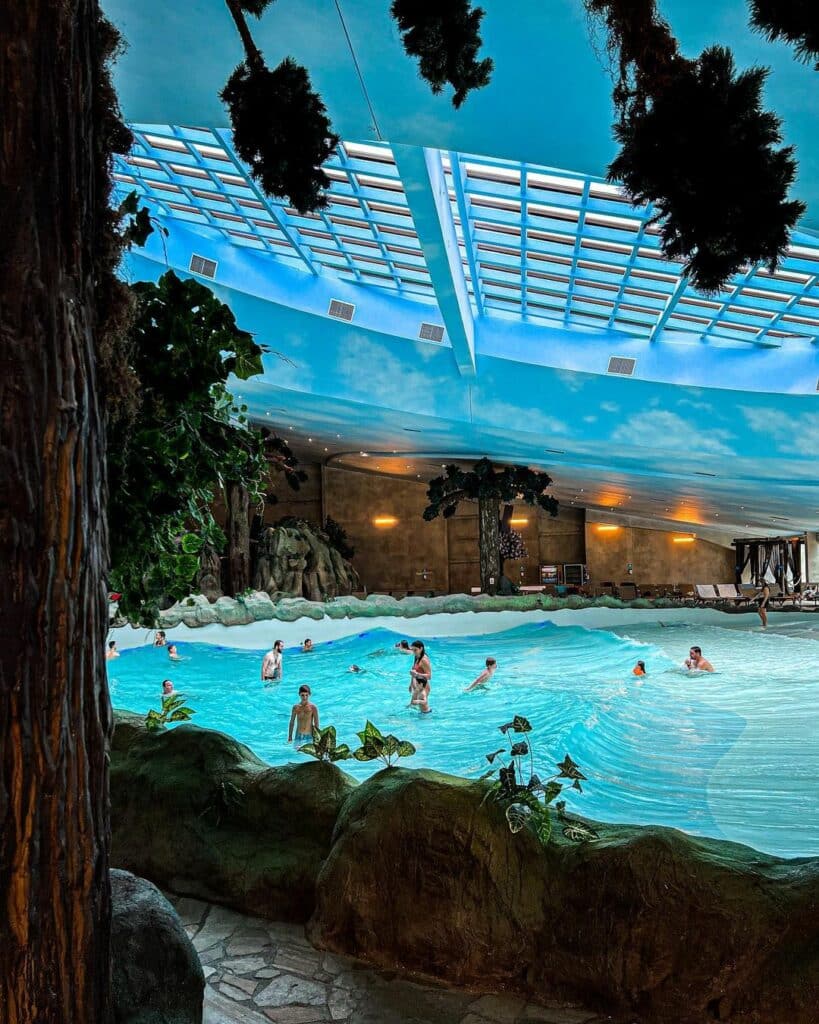 A imagem mostra o Acquamotion, um parque aquático coberto em Gramado, RS. No centro da foto, há uma grande piscina de ondas onde várias pessoas estão se divertindo. O teto do parque é transparente, permitindo a entrada de luz natural, e tem um design que simula um céu azul com nuvens brancas, aumentando a sensação de estar ao ar livre. A área ao redor da piscina é decorada com plantas artificiais e árvores, criando um ambiente de floresta tropical. Ao fundo, há espreguiçadeiras para relaxamento, e a iluminação suave do ambiente contribui para a atmosfera acolhedora e divertida do local. A foto foi reproduzida do Instagram.