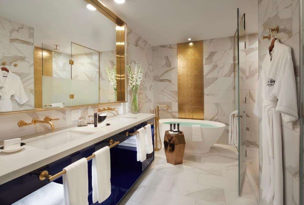 Banheiro luxuoso do Hard Rock Hotel com duas pias, uma banheira independente, e uma decoração em mármore e dourado. Há roupões pendurados na parede e flores brancas sobre a bancada. Este é um dos hotéis em Nova York.