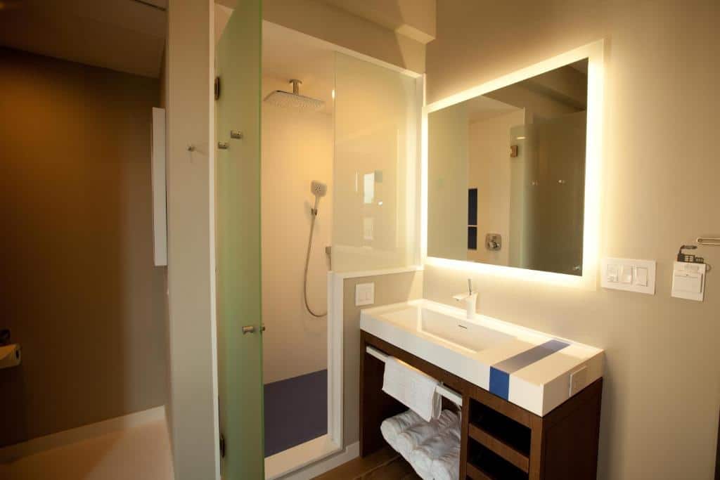 Banheiro moderno de hotel com um chuveiro fechado por uma porta de vidro e uma pia com espelho iluminado. O design é contemporâneo, com uma combinação de cores suaves.