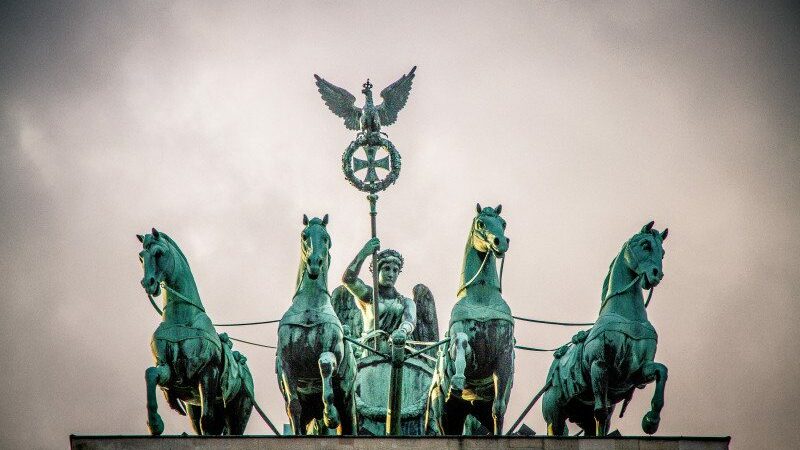 Imagem do monumento do Portão de Brandemburgo durante o dia com quatro cavalos e um homem em um carruagem. Representa o que fazer em Berlim.