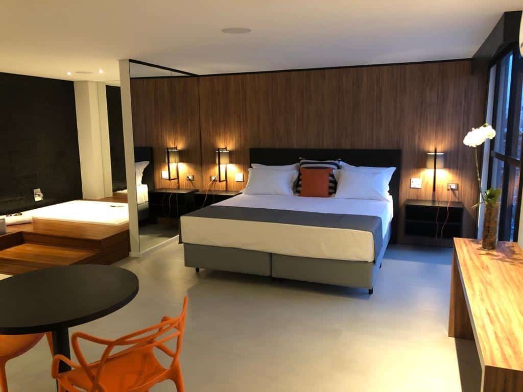 Quarto do Best Guest Hotel Expo Anhembi. Uma cama de casal no centro, uma mesa, uma banheira de hidromassagem e uma parede de espelho do lado esquerdo. Do lado direito uma parede de vidro. Imagem para ilustrar post sobre hotéis perto do Expo Center Norte.