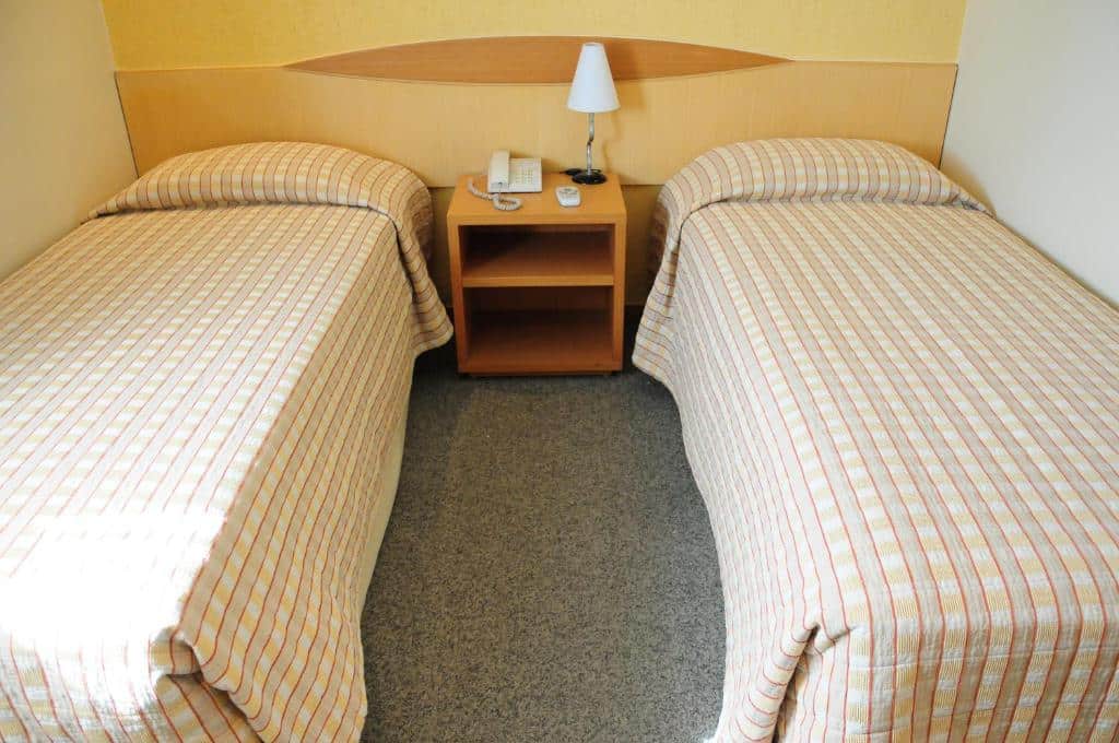 Quarto da Brasília Santana Gold Flat. Uma cama de solteiro de cada lado e uma cômoda com um abajur no centro. Imagem para ilustrar post sobre hotéis perto do Expo Center Norte.