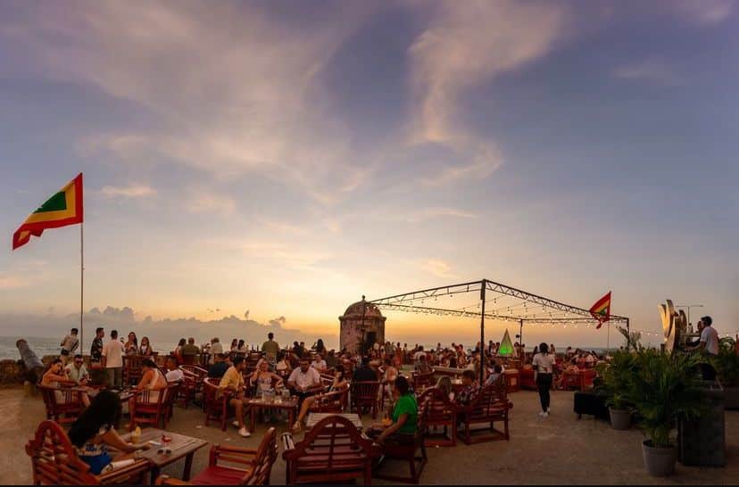 A imagem mostra um bar ao ar livre, o Café del Mar, localizado em Cartagena, durante o pôr do sol. Várias mesas e cadeiras de madeira estão dispostas em um terraço amplo, onde pessoas relaxam e desfrutam de suas bebidas e refeições. No fundo, a linha do horizonte do oceano e um céu pintado com tons de rosa, laranja e azul proporcionam um cenário deslumbrante. Duas bandeiras coloridas estão hasteadas, e luzes penduradas adicionam um toque acolhedor e festivo ao ambiente.