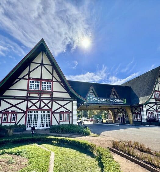 A imagem retrata a entrada da cidade de Campos do Jordão, em São Paulo. No centro, entre dois edifícios de arquitetura alpina, há um telhado com uma placa que diz o nome da cidade. O céu está ensolarado e com poucas nuvens.